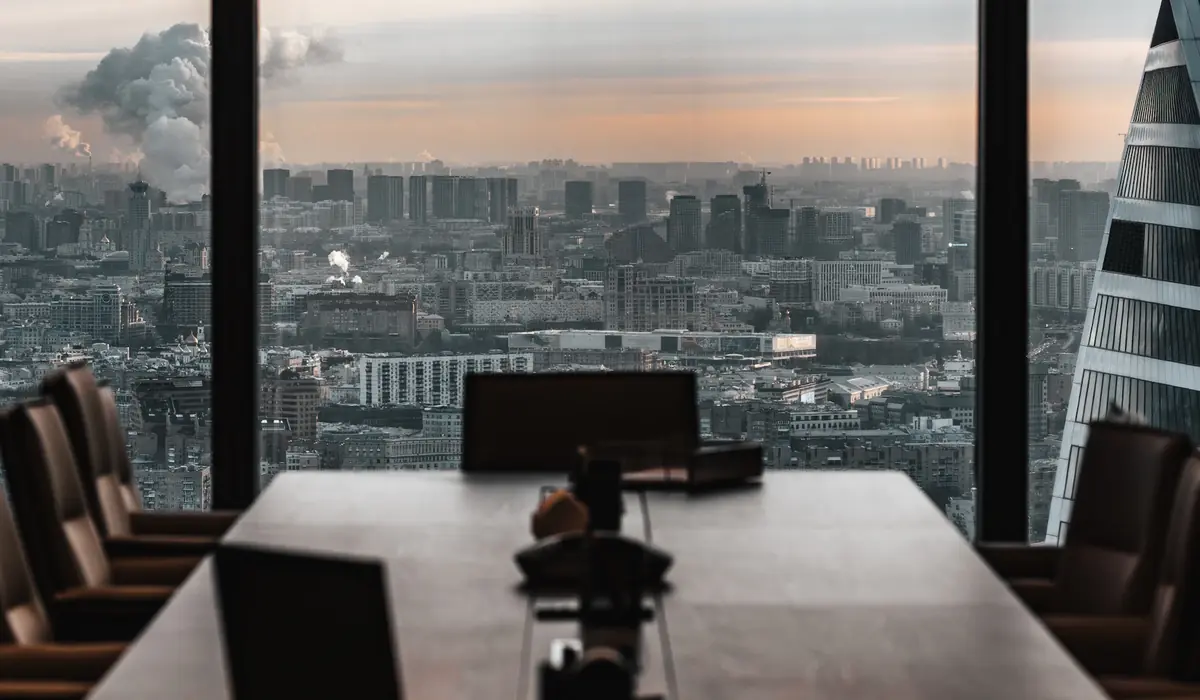 An empty conference table in front of a tall window overlooking a city at dusk